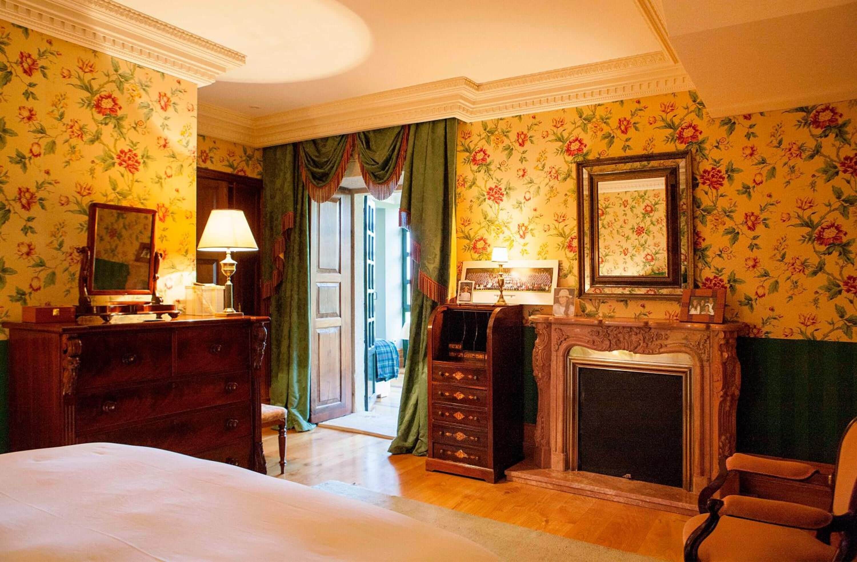 Een klassieke kamer met bloemenbehang, een open haard, een bed, een kaptafel en een spiegel.