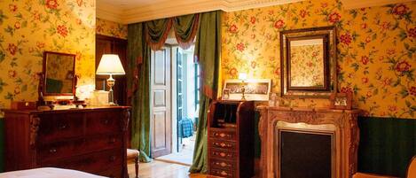 Een klassieke kamer met bloemenbehang, een open haard, een bed, een kaptafel en een spiegel.
