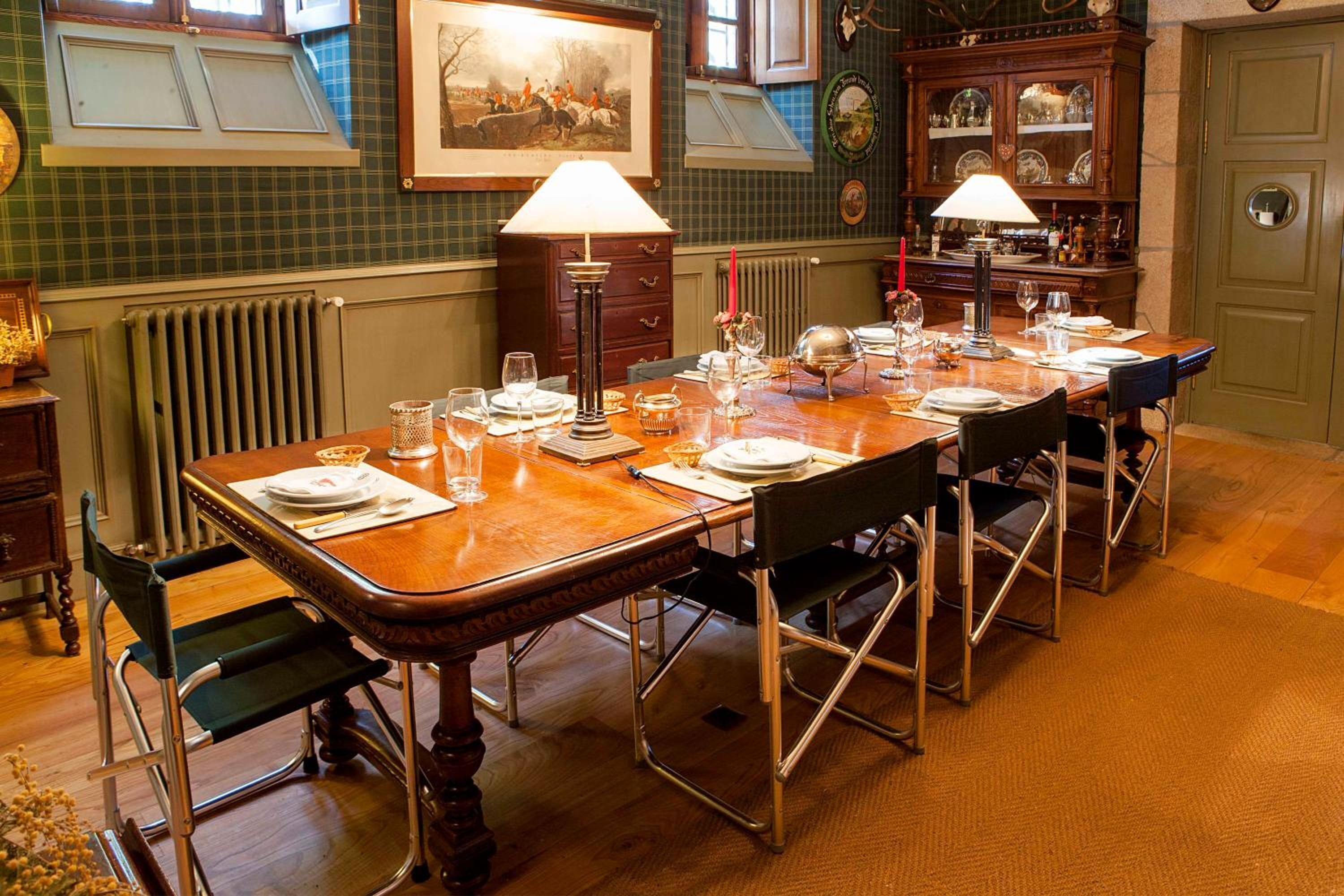 Een eetkamer met een lange houten tafel, gedekt voor zes personen, met aan weerszijden lampen, een radiator en een kast met glazen.
