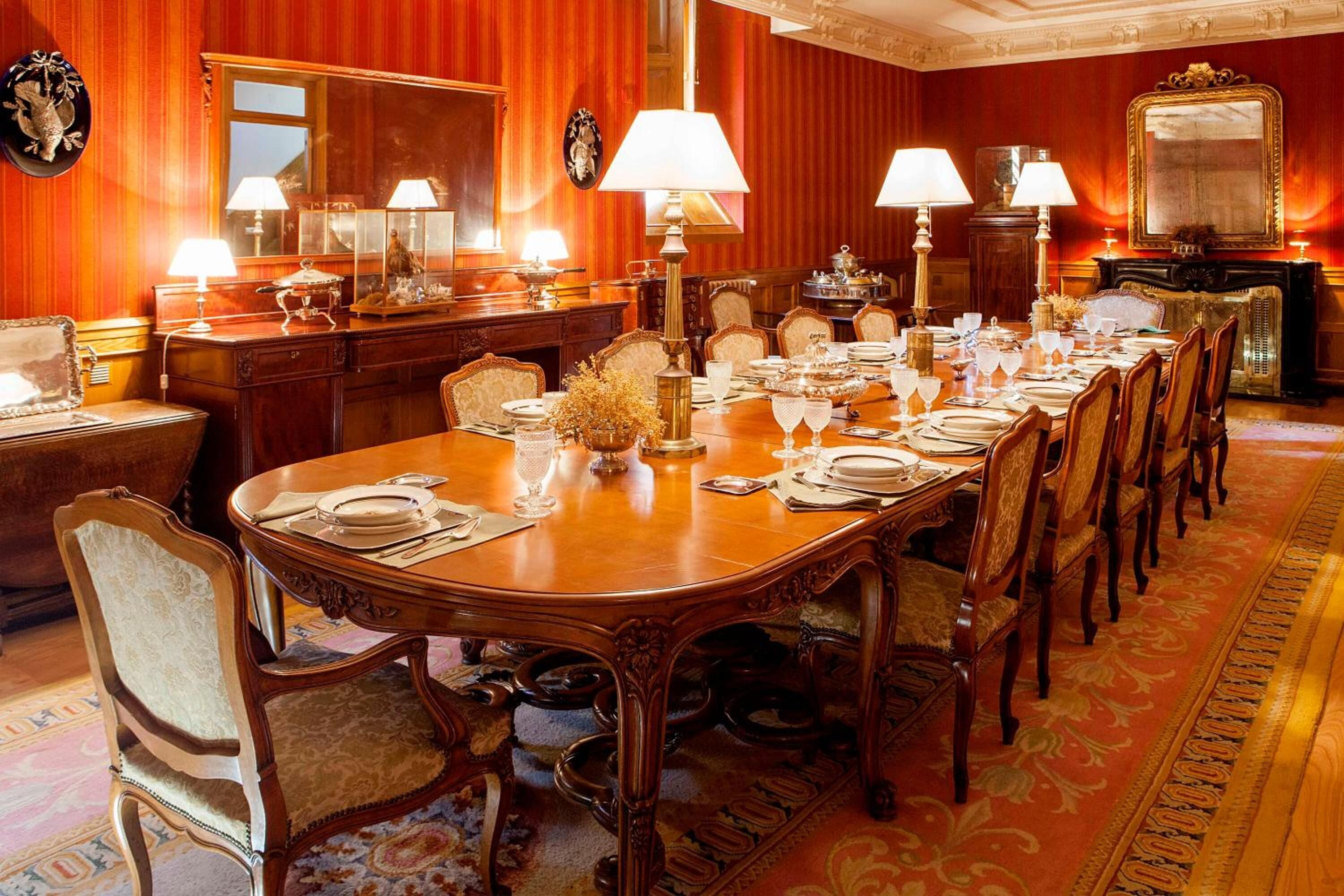 Uma sala de jantar formal com uma longa mesa de madeira posta para uma refeição, rodeada por cadeiras ricamente decoradas.