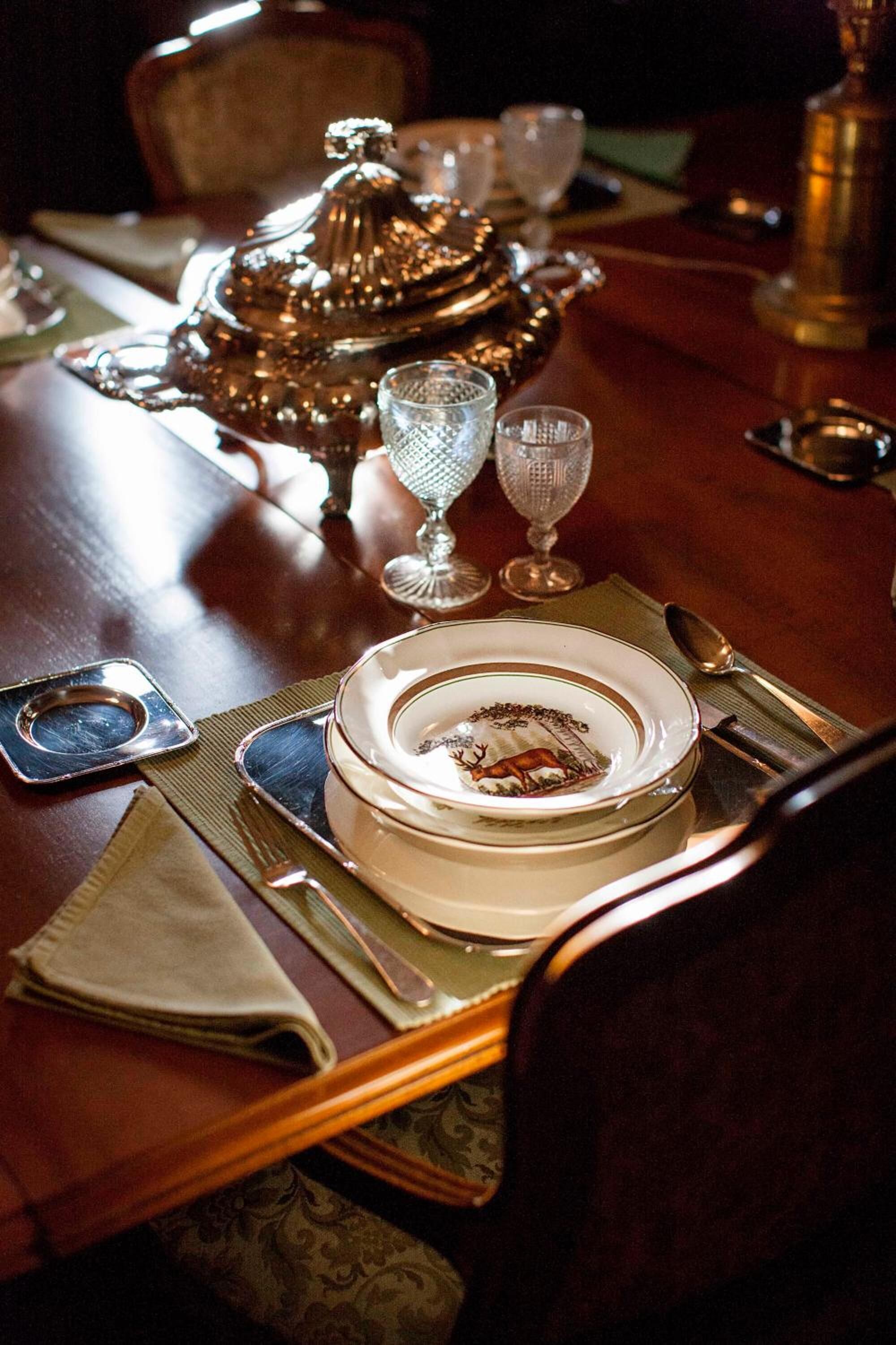 Una mesa de comedor formal decorada con una tureen de plata, copas de cristal y un plato con diseño de ciervo.