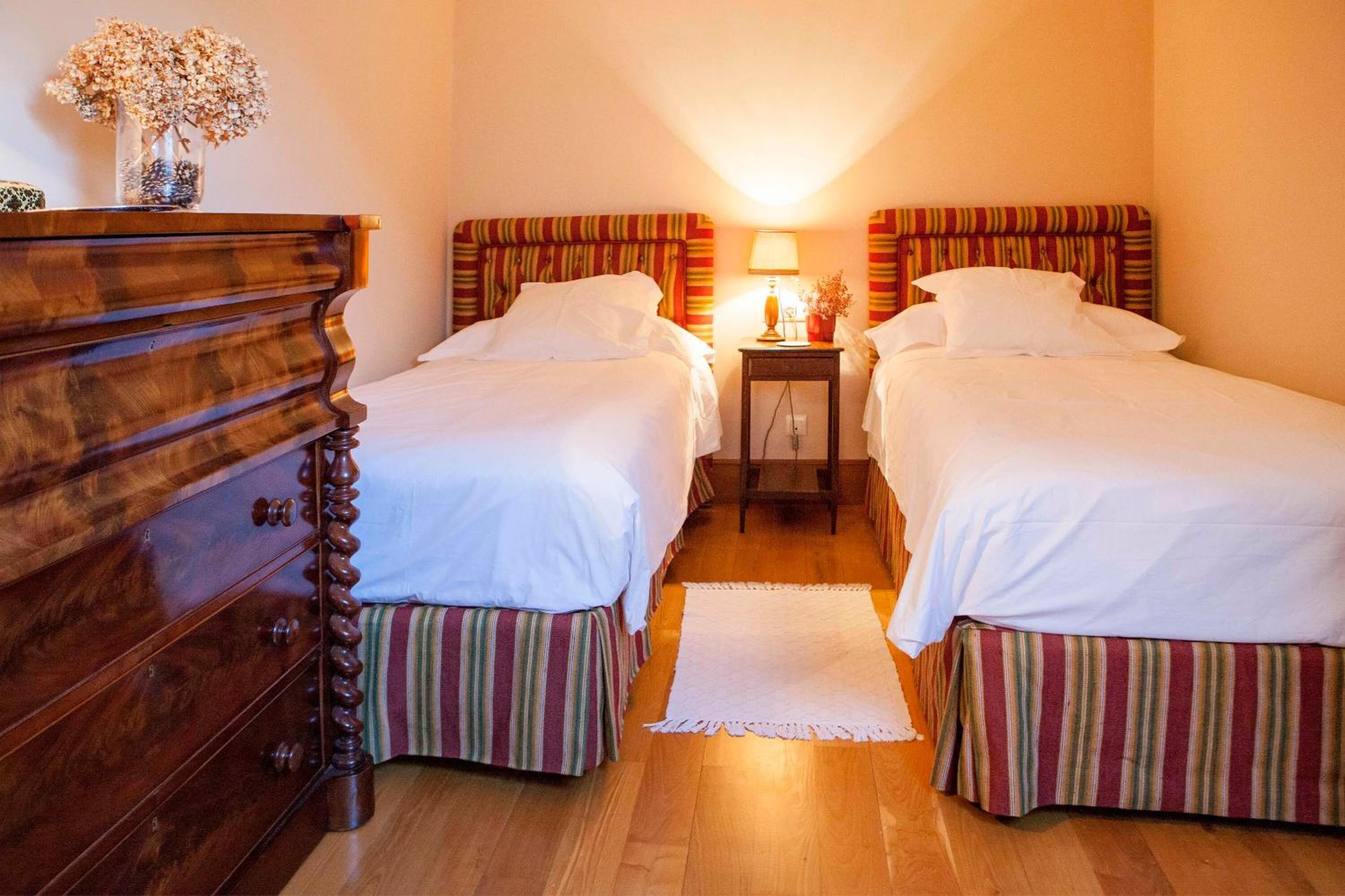 Hotellihuone, jossa on kaksi sänkyä, puinen lipasto, yöpöytä lampun kanssa ja pieni pöytä, jossa on maljakko täynnä kukkia.
