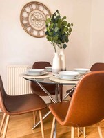 Un comedor con una mesa redonda de vidrio, sillas de cuero marrón, platos blancos y un reloj de pared decorativo.