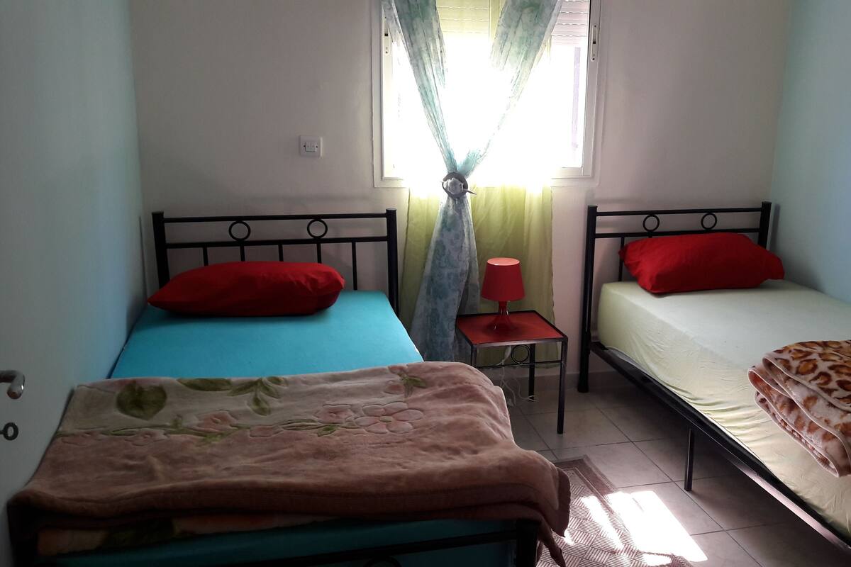 A room with two beds, each with a red pillow, a small table with a red lamp, and a window with sheer curtains.