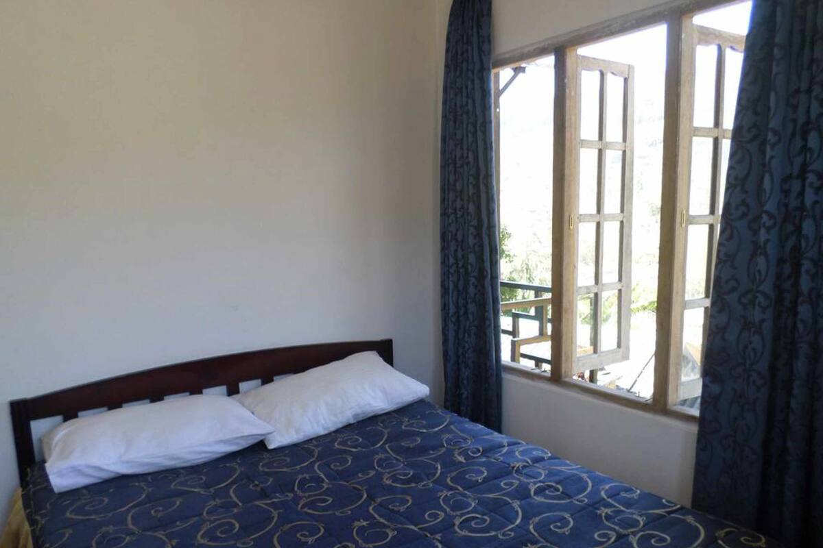 A bed with a dark blue patterned bedspread and two white pillows in a room with a large window and curtains.