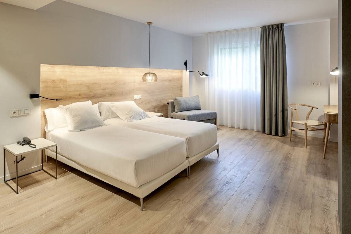 A modern hotel room with a wooden headboard, two beds, a desk, and a chair.