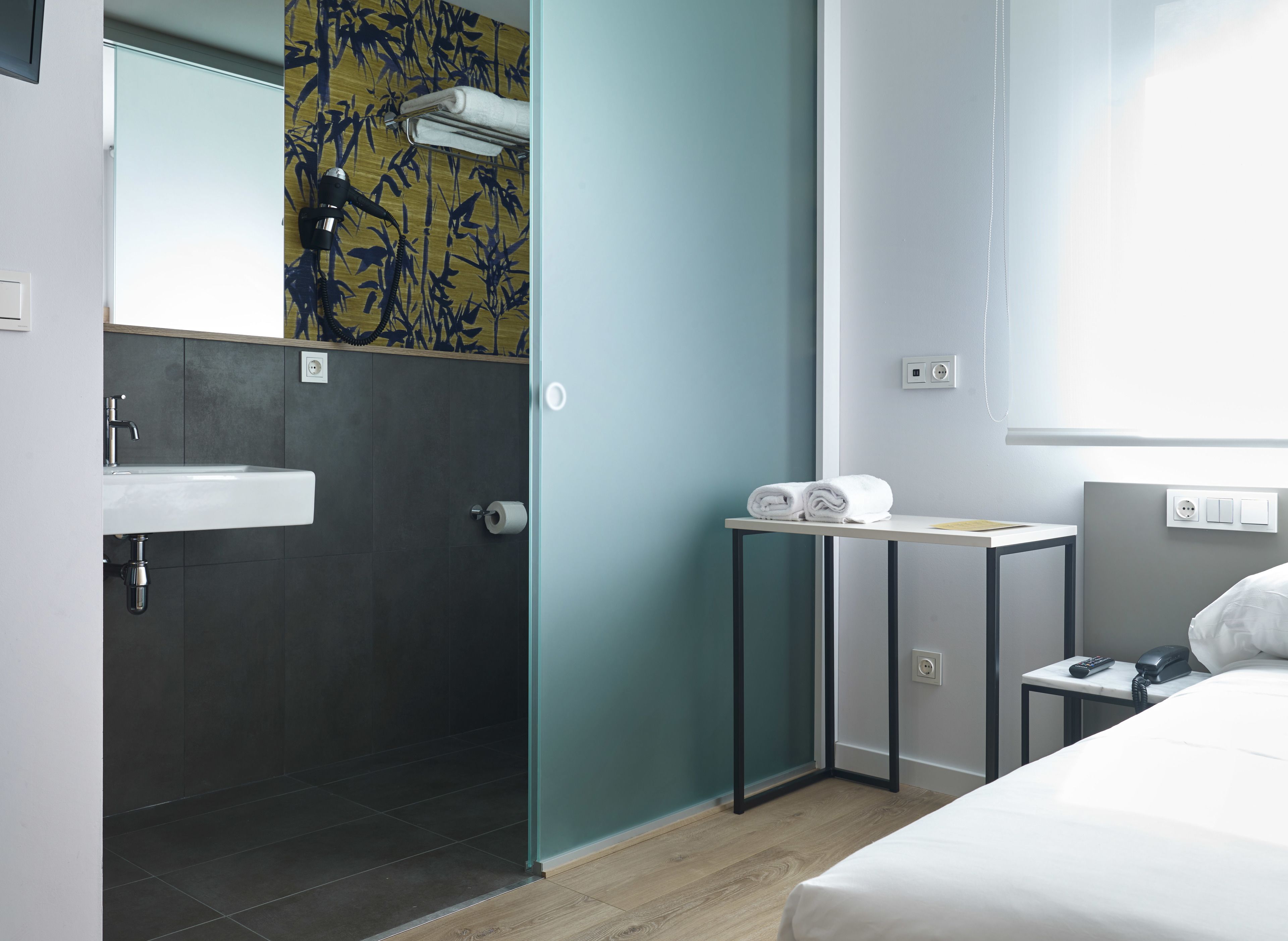 Una habitación de hotel moderna con baño, una cama, una mesita de noche con teléfono y un perchero para toallas.