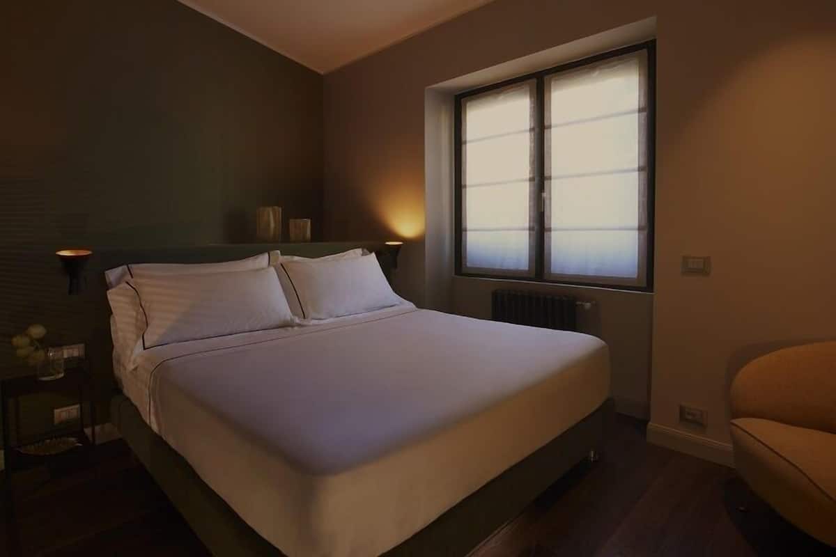 A bedroom with a bed, a window with blinds, a nightstand with a lamp, and a chair.