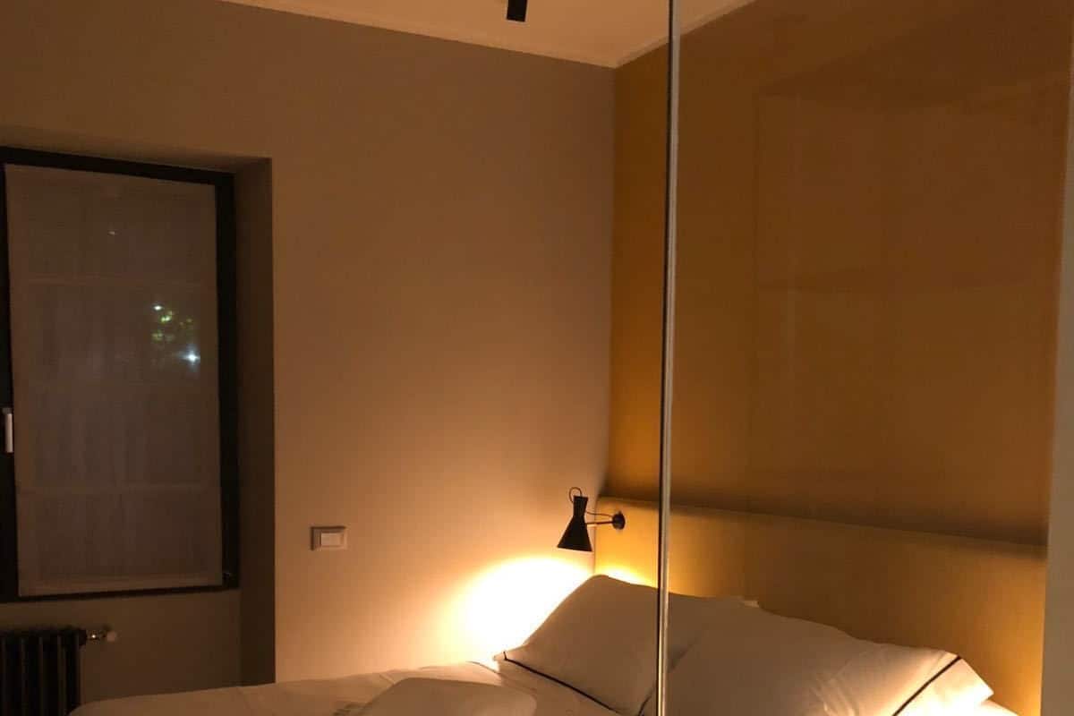 A hotel room with a bed, a nightstand, a wall-mounted lamp, and a window with a view of the night sky.