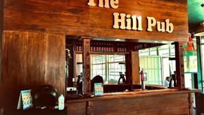 El Hill Pub con interior de madera, mostrador de bar, taburetes y un panel de menú visible.