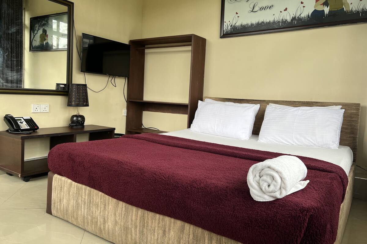 A hotel room with a bed, a desk, a telephone, a television, and a painting on the wall.