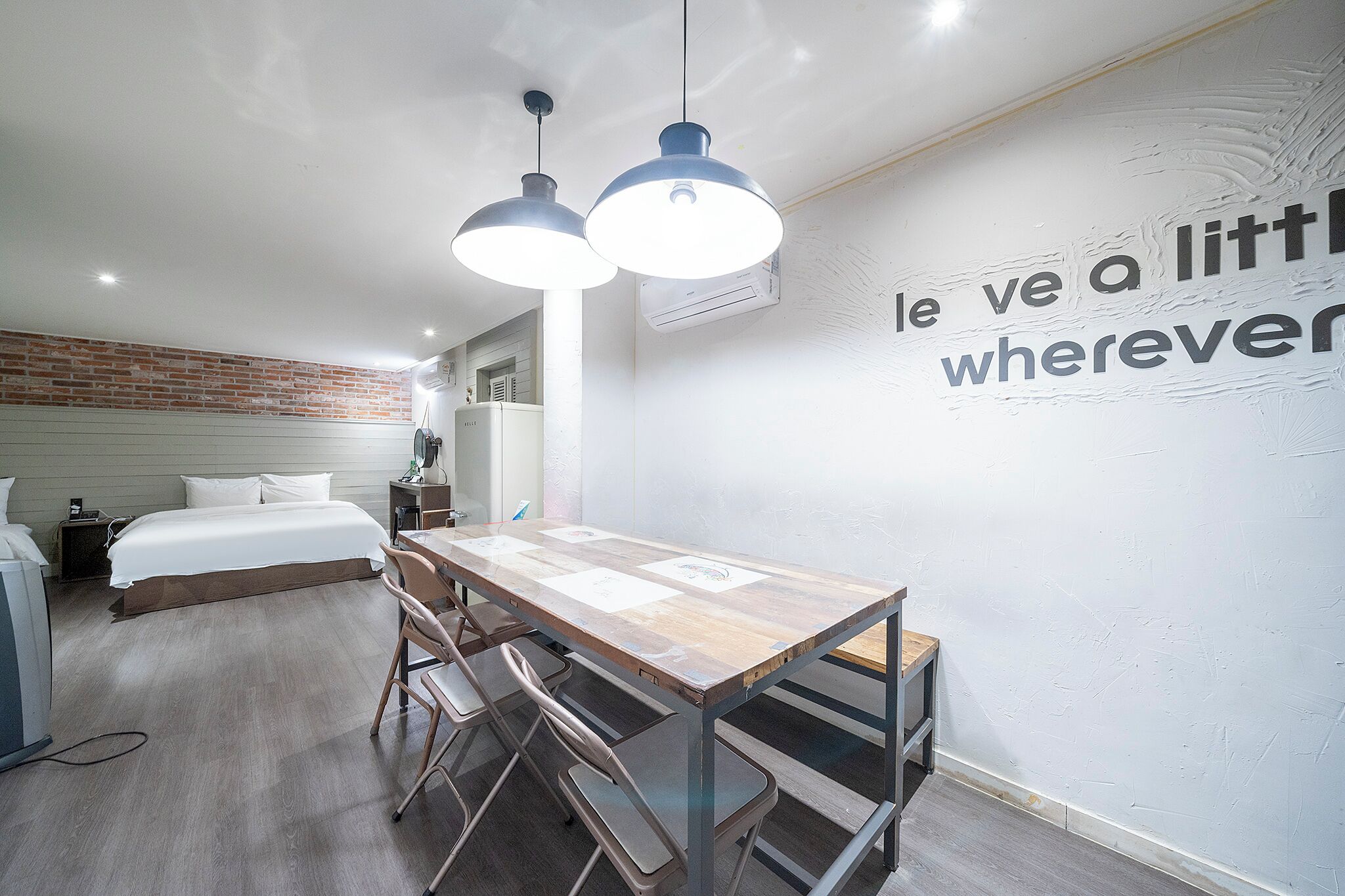 Una habitación de hotel moderna con cama, mesita de noche y escritorio con sillas. Cuenta con un equipo de aire acondicionado de pared y una lámpara de techo.