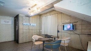 Un moderno vestíbulo de hotel con una pared de concreto, un letrero de neón que dice "HOTEL YOJA" y una pantalla de televisión mostrando una pregunta.