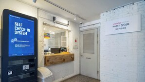 Un quiosco de auto-registro, un mostrador de recepción y un cartel que dice 'Yanoljo Hotel Yaja' en una habitación con paredes de ladrillo blanco y piso de baldosas.