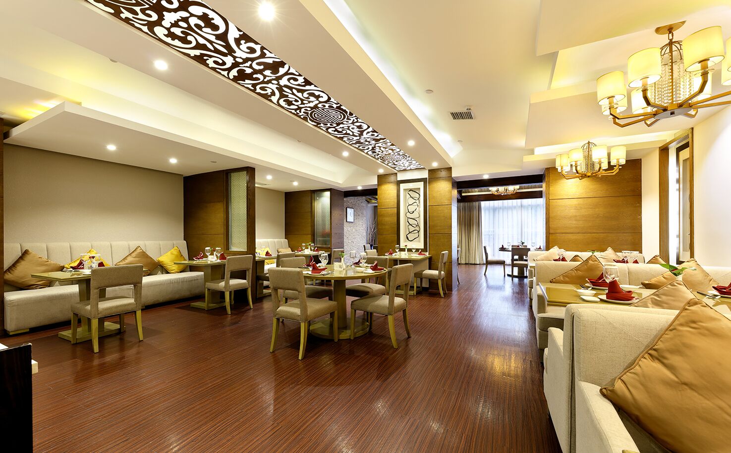 A spacious hotel dining area with multiple tables and chairs, a decorative ceiling panel, and a chandelier.