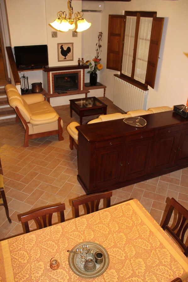 A living room with a fireplace, a television, and a dining area with a table set for two. - Agriturismo Cantagalli (San Quirico d'Orcia)