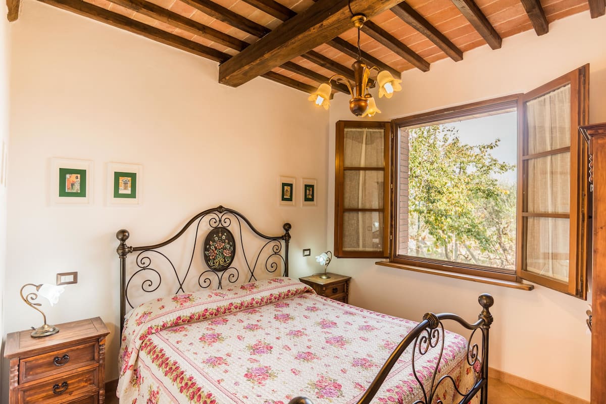 A bedroom with a bed, wooden headboard, a nightstand, a lamp, and a window with a view of trees.