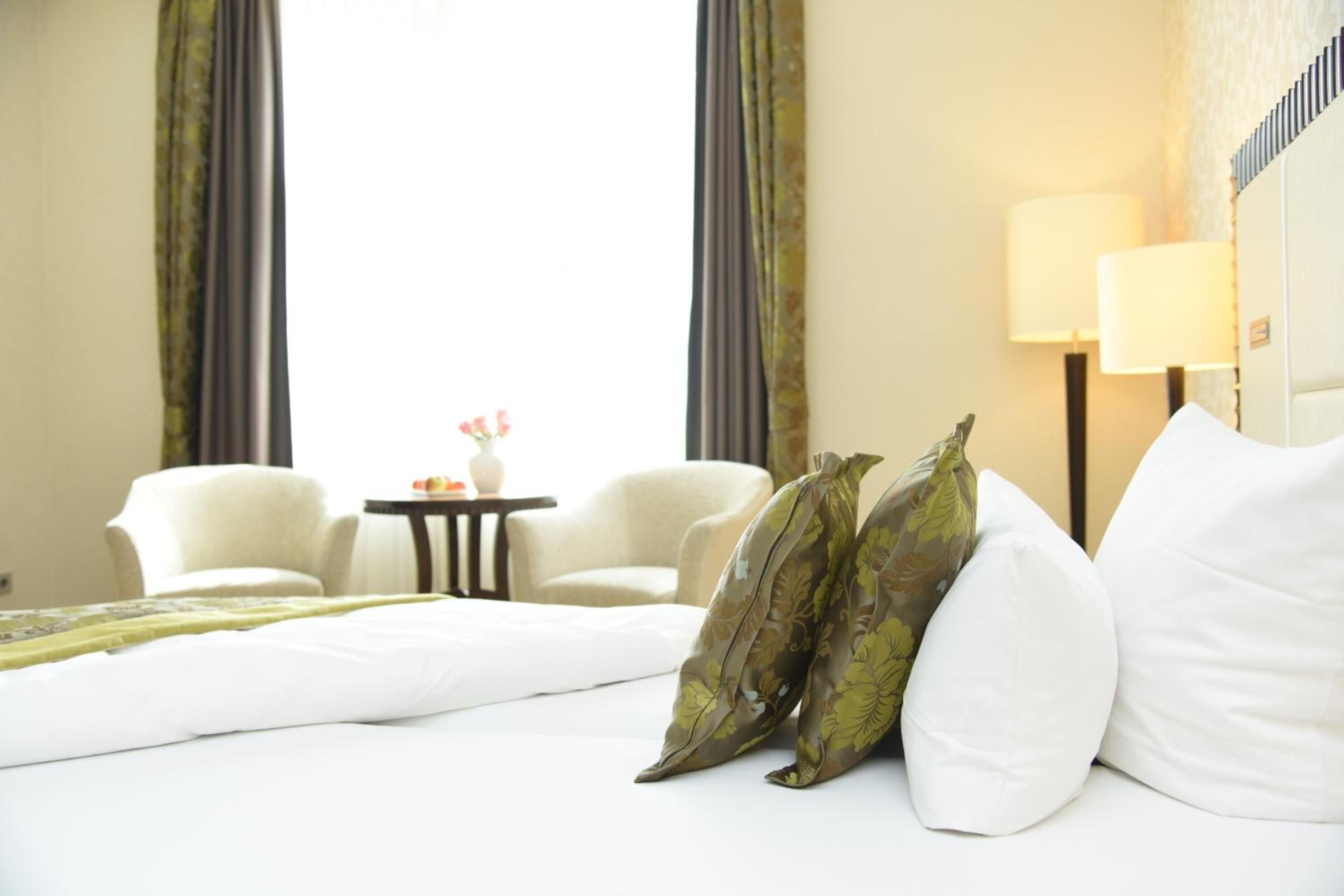 Ein Hotelzimmer mit einem Bett, zwei weißen Stühlen, einem kleinen Tisch mit Vase und Blumen sowie einer Lampe.