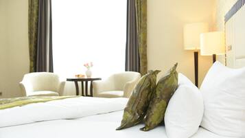 Ein Hotelzimmer mit einem Bett, zwei weißen Stühlen, einem kleinen Tisch mit Vase und Blumen sowie einer Lampe.
