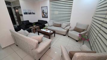 A living room with beige sofas, a coffee table, and a potted plant. There is a painting on the wall and a window with blinds.