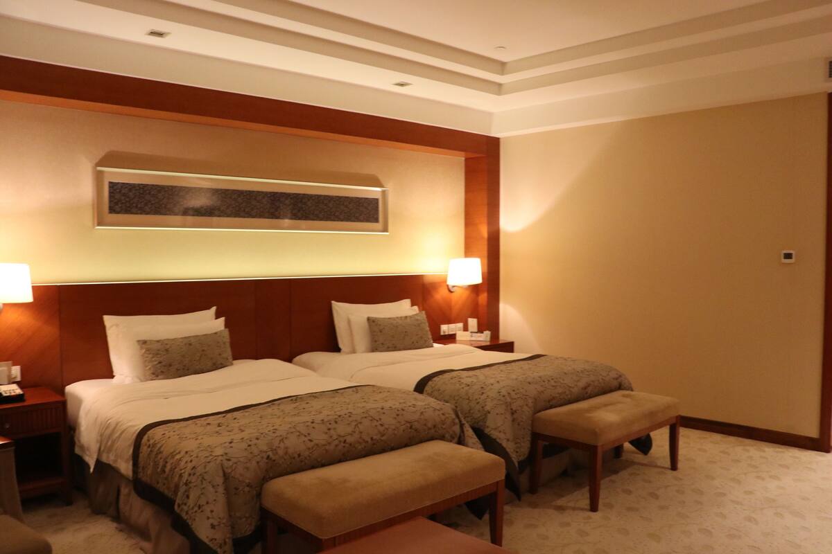 Ein Hotelzimmer mit zwei Betten, einem Nachttisch mit Lampe, einer Bank und einem Wandbild.