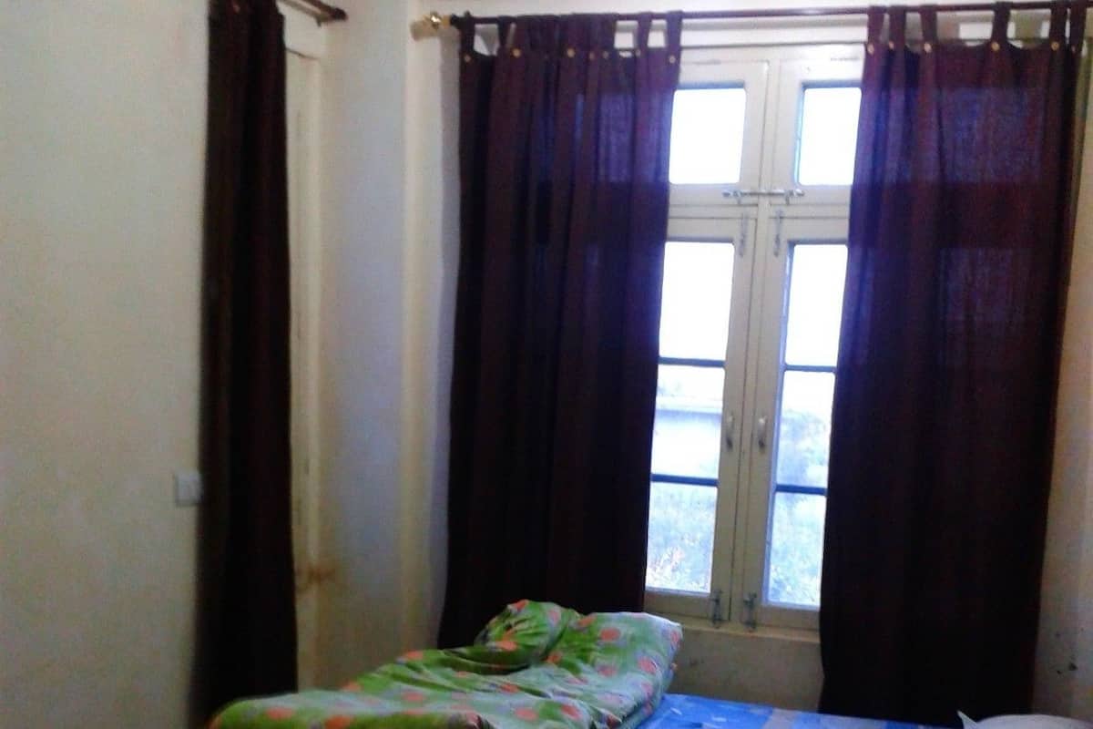 A single bed with a blue and white checkered bedspread, a green blanket with a floral pattern, and two pillows. The room has a window with dark curtains and a view of trees outside.