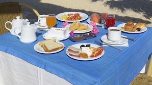 Une table installée sur la plage, avec un assortiment de produits pour le petit-déjeuner, notamment des toasts, des œufs et des fruits, le tout mis en valeur par un ciel bleu et dégagé.
