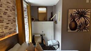 Et hotelværelse med en væg i murstensmønster, en seng med hvide hør sengetøj, mønstrede puder, et spejl, et billede af planter og en dør med et håndklædestativ.