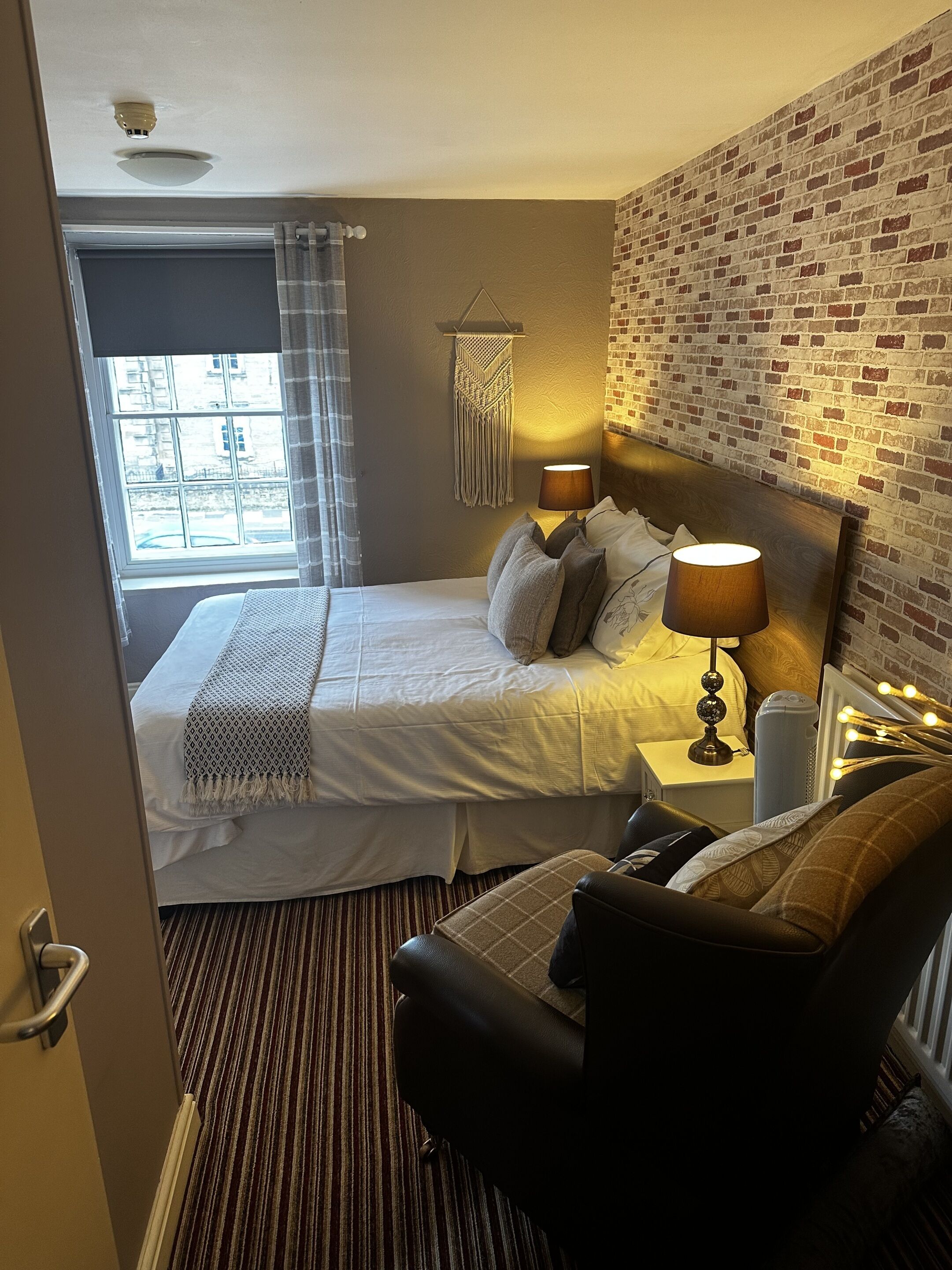 A hotel room with a bed, a chair, a bedside table with a lamp, a macrame wall hanging, and a brick-patterned wall.