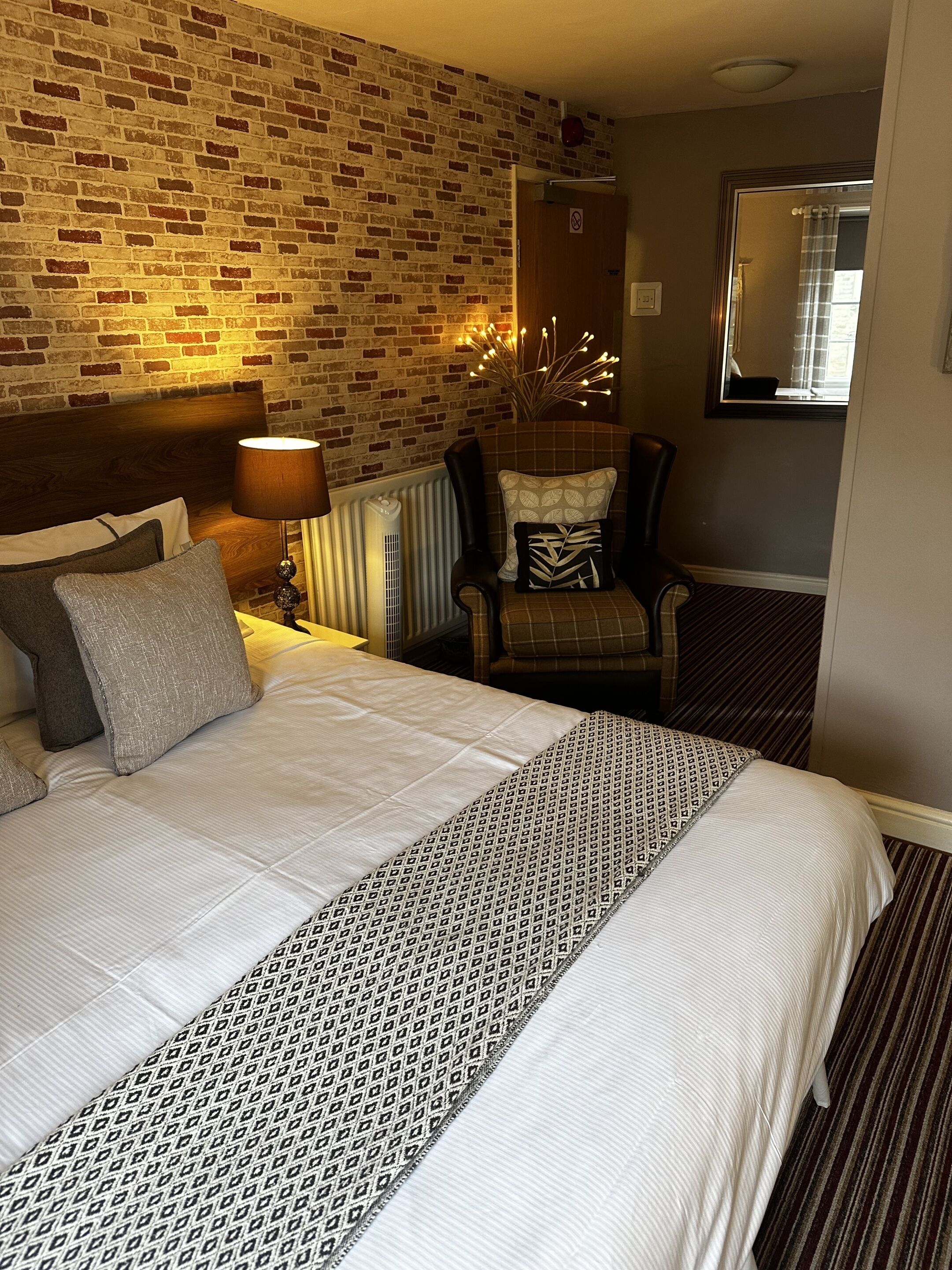 Een hotelkamer met een metselwerk muur, een groot bed met beddengoed met een patroon, een stoel met een kussen, een nachtkastje met een lamp en een spiegel aan de muur.
