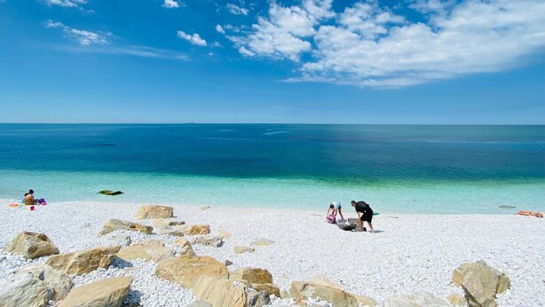 A beach with white pebbles and large rocks, clear turquoise water, and a few people. - Villino Ermione (Pisa)