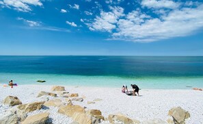 A beach with white pebbles and large rocks, clear turquoise water, and a few people. - Villino Ermione (Pisa)