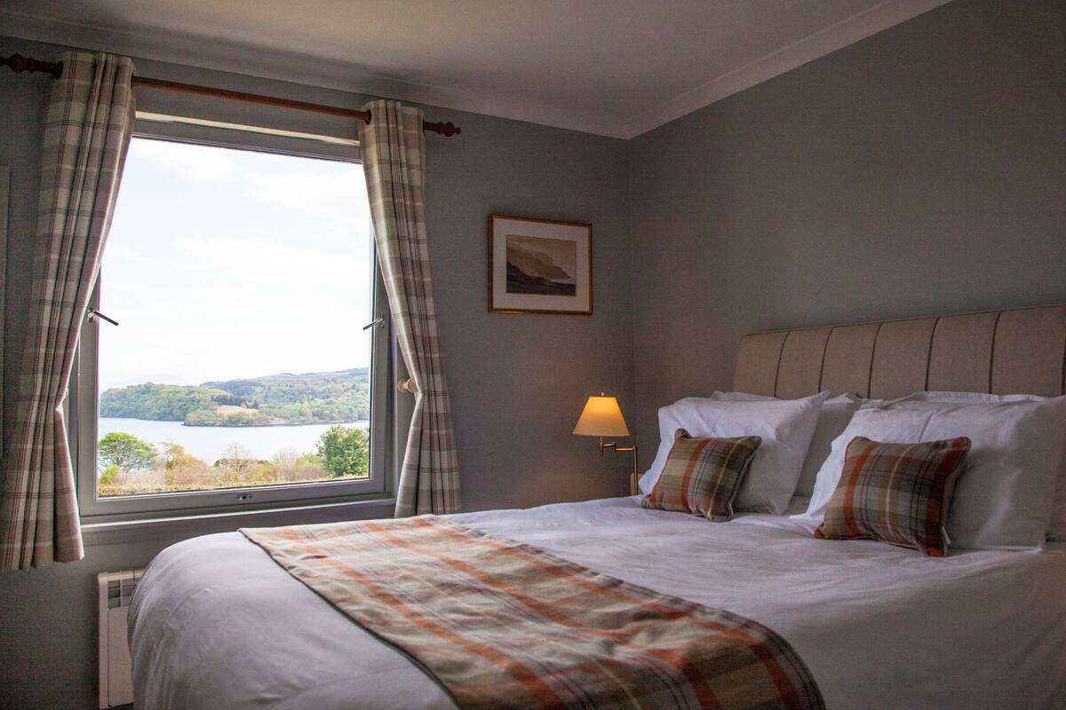 A bedroom with a large bed, a window with curtains, a bedside lamp, and a framed picture on the wall.