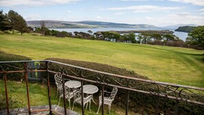 A balcony with wrought iron railings and white metal chairs overlooking a grassy landscape with trees and a body of water in the distance. - Fairways Lodge B&B (Isle of Mull)