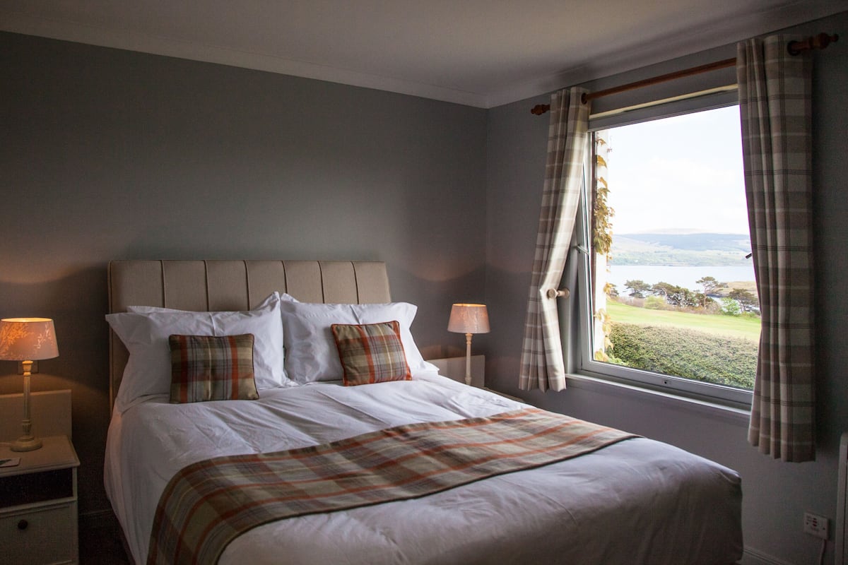 A neatly made bed with a plaid throw blanket, two bedside lamps, and a view of a lake through a window with curtains.