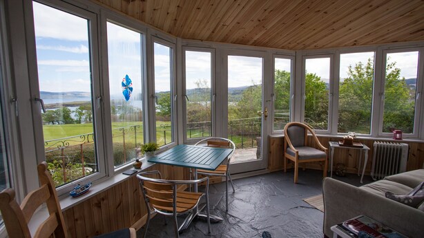 A room with a wooden ceiling, large windows, a table with chairs, a sofa, and a radiator. - Fairways Lodge B&B (Isle of Mull)