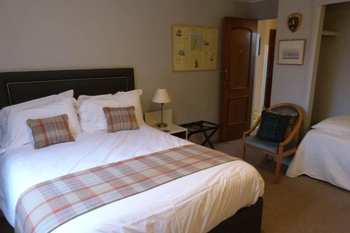 A neatly made bed with a plaid bedspread, a wooden nightstand with a lamp, a wooden chair, and a door with a coat hanging on it.
