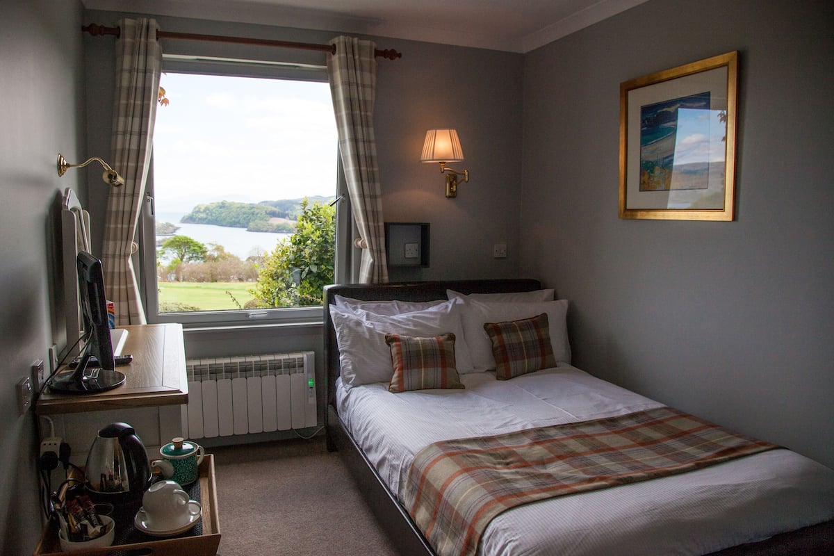 A bedroom with a bed, a bedside table with a lamp, a window with curtains, and a painting on the wall.