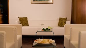 A room with a white sofa, two green patterned pillows, a glass coffee table with a floral arrangement, and a plate of food.
