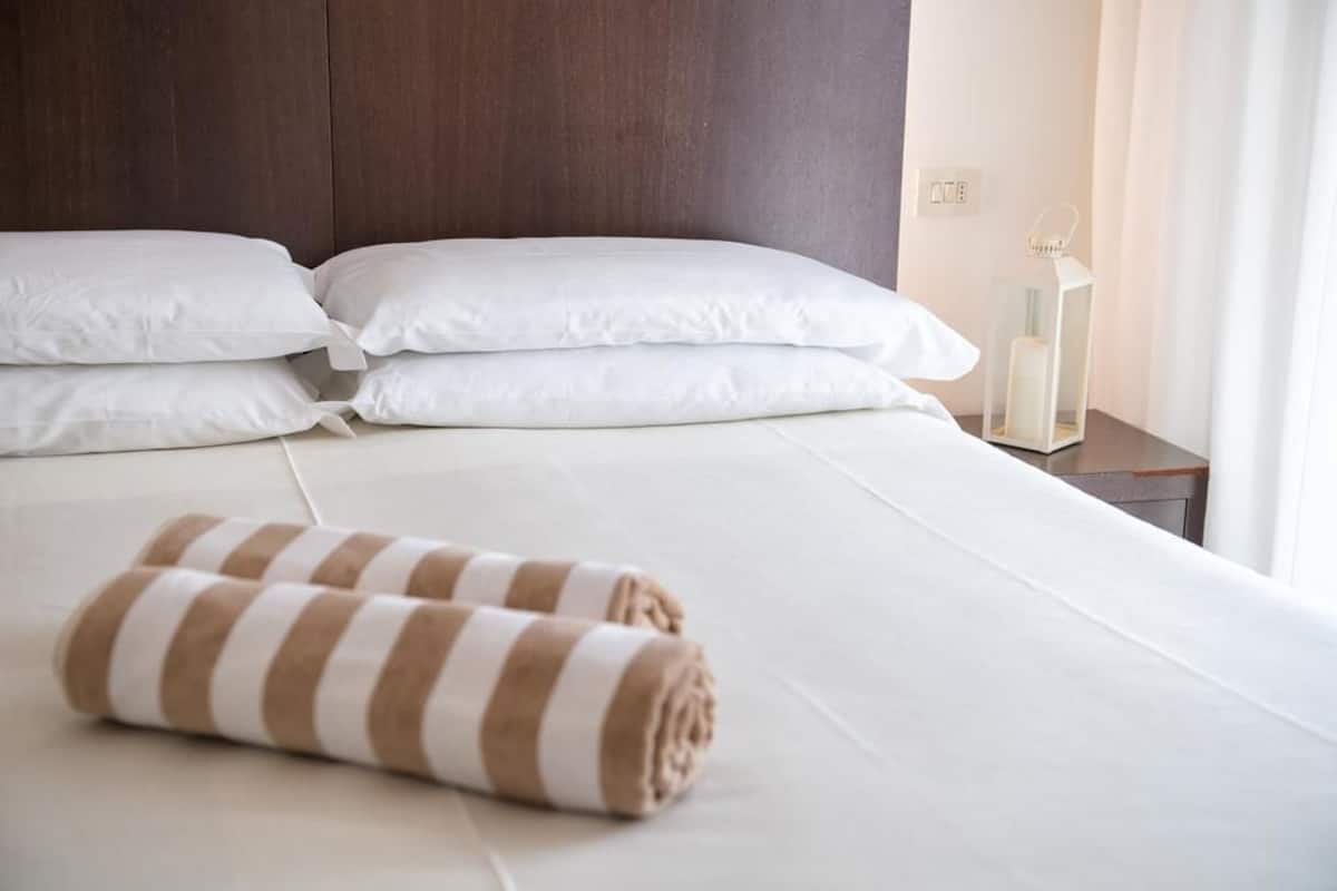 Un lit bien fait, avec du linge de lit blanc et une serviette à rayures, une tête de lit en bois et une table de chevet surmontée d’une lampe blanche.