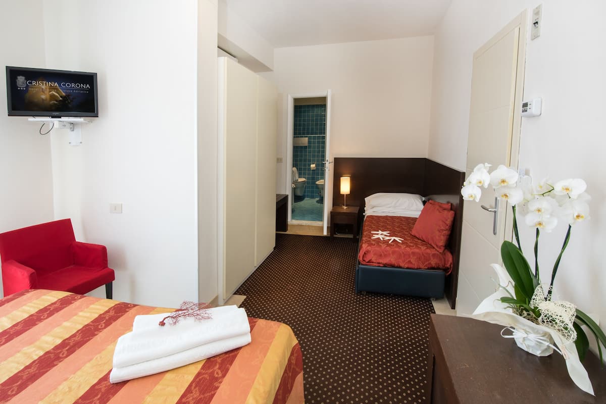 Une chambre d’hôtel avec un téléviseur fixé au mur, un fauteuil rouge, un lit recouvert d’une courtepointe à rayures, une table de chevet ornée d’un vase de fleurs blanches et une salle de bain visible à travers une porte ouverte.