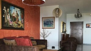 A living room with a brown sectional sofa, a yellow ottoman, a white pendant light, and a chandelier. There are paintings on the walls and a door in the background. - Nice view Apartment (Chnaneir)