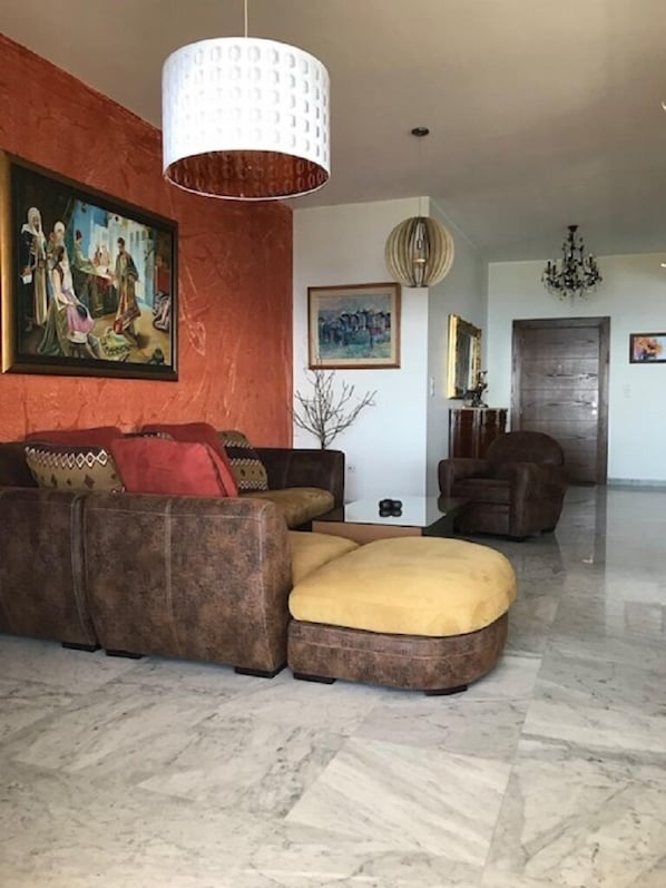 A living room with a brown sectional sofa, a yellow ottoman, a white pendant light, and a chandelier. There are paintings on the walls and a door in the background.