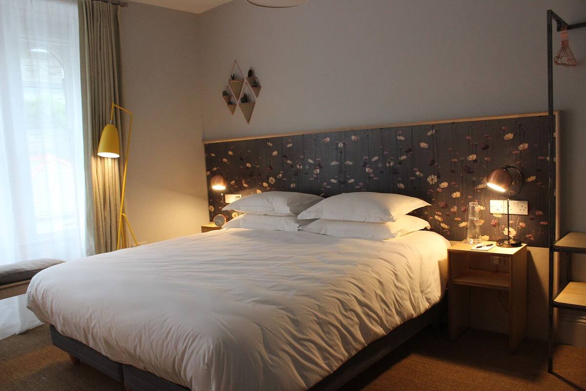 A neatly made bed with white linens, a bedside lamp, a clock, and a floral-patterned headboard.