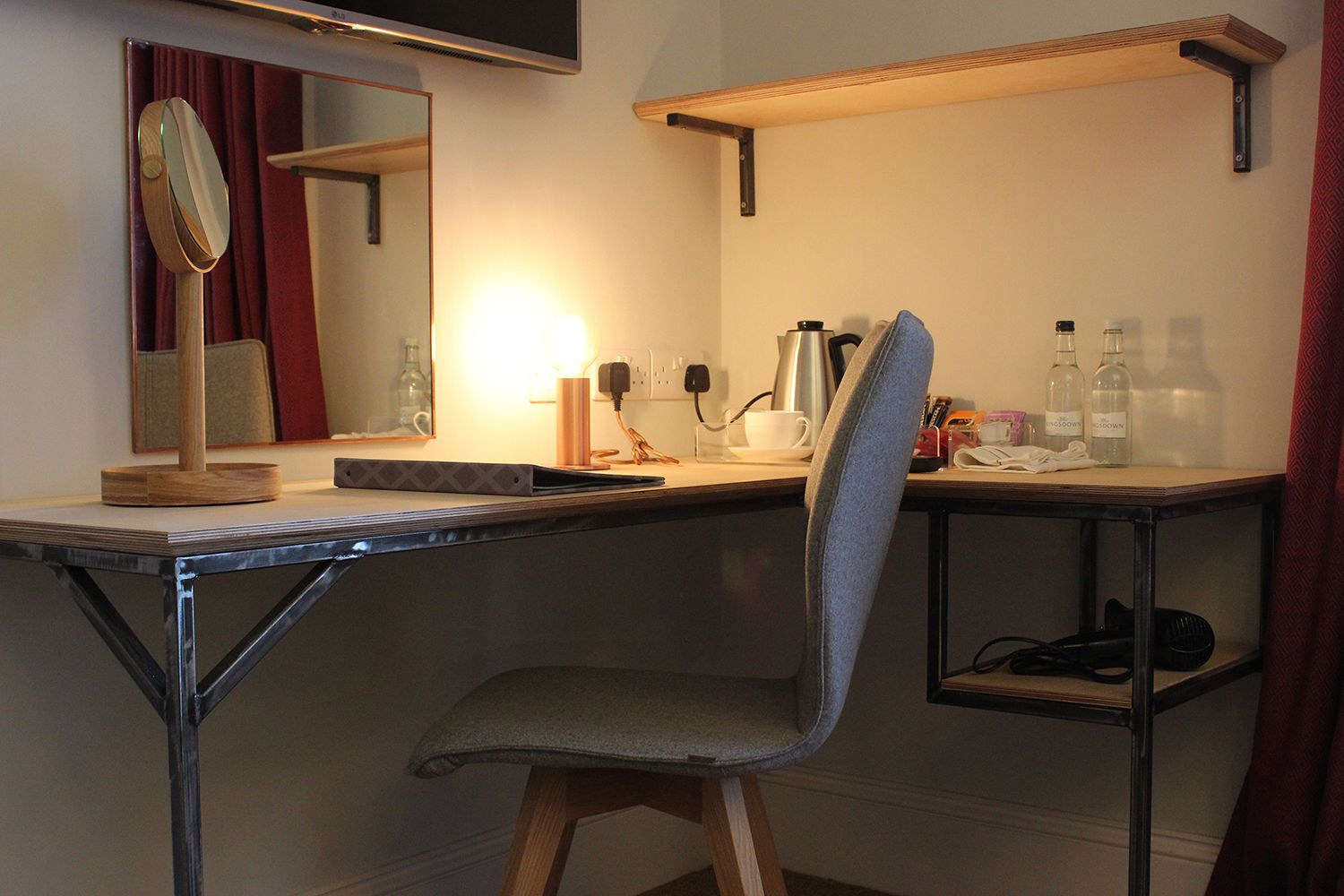 Ein kleines Hotelzimmer mit Schreibtisch, Stuhl, Spiegel und Ablagefläche für Toilettenartikel.