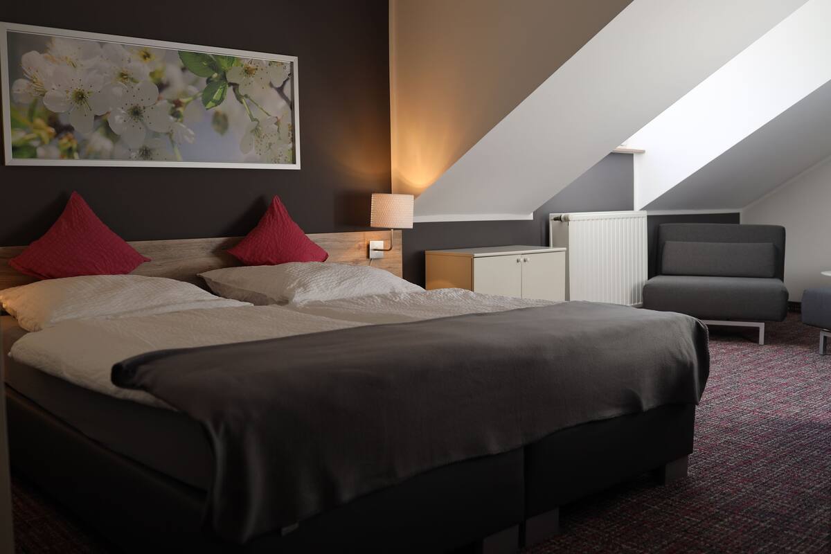 A modern hotel room with a large bed, two red pillows, a nightstand with a lamp, a gray armchair, and a large window with natural light.