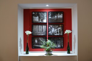 Una ventana con cuatro fotografías blanco y negro sobre una pared roja, un jarrón verde con flores blancas y dos botellas verdes a cada lado.