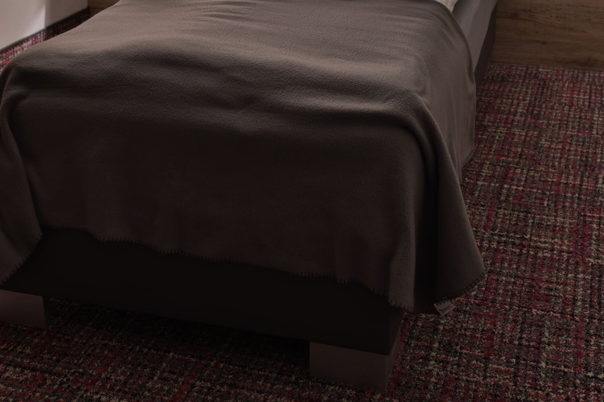A neatly made bed with a dark blanket, a wooden headboard, a bedside table with a lamp, and a framed picture on the wall.