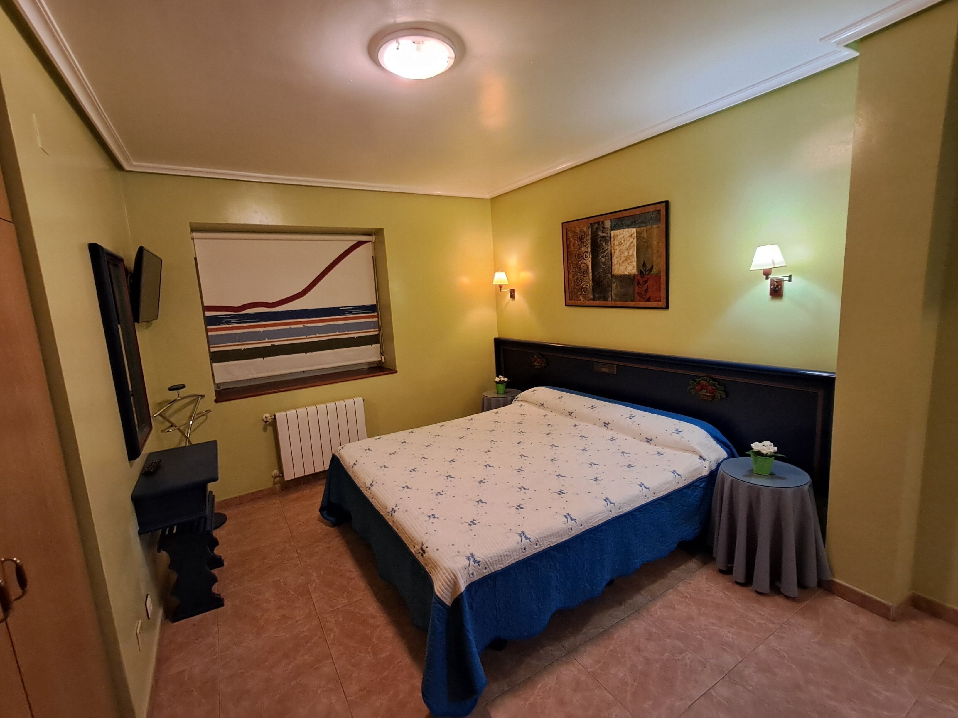 A bedroom with a bed, a wall-mounted TV, a radiator, a lamp, and a painting.
