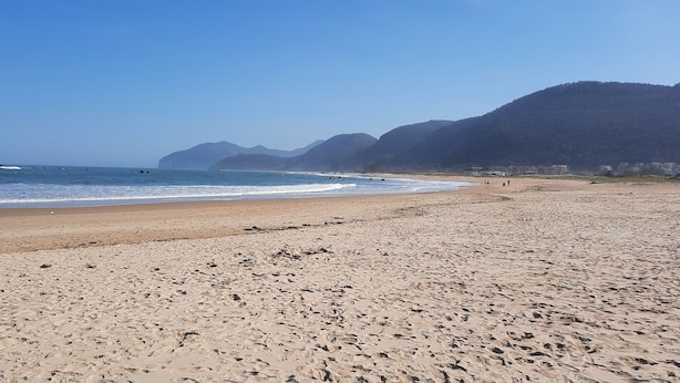 A wide sandy beach with footprints, calm sea, and mountains in the background.